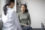 A young female patient sits casually with her doctor as they discuss her mental health. She is seated in a chair in front of her doctor as they talk about her needs. The doctor is wearing a white lab coat and has her back to the camera as the two discuss possible plans of care to navigate the woman's struggles.